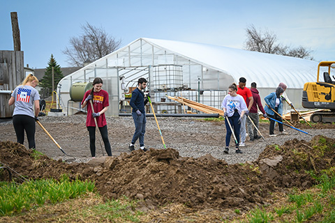 Students with rakes standing on gravel near dirt piles in front of a greenhouse.